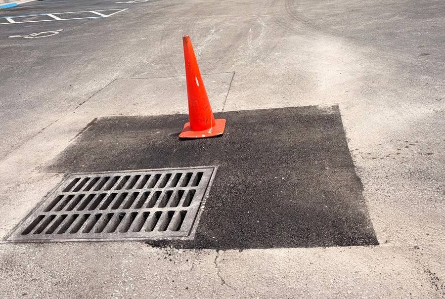 Parche de asfalto nuevo alrededor de la rejilla de un tragante en un estacionamiento comercial, señalizado con un cono